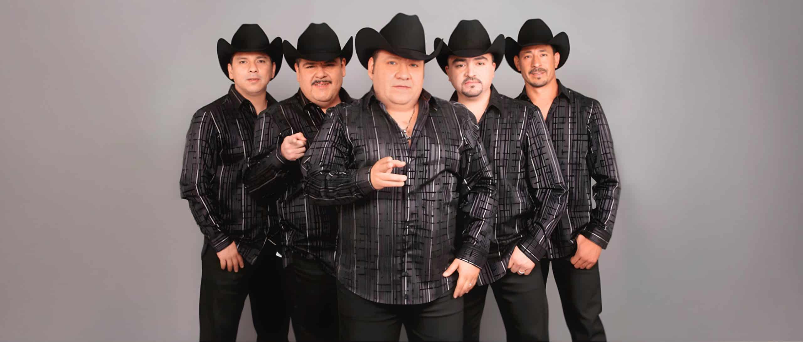 Grupo Pesado posando con vestimenta negra y sombreros norteños, formación completa de la banda lista para show en vivo