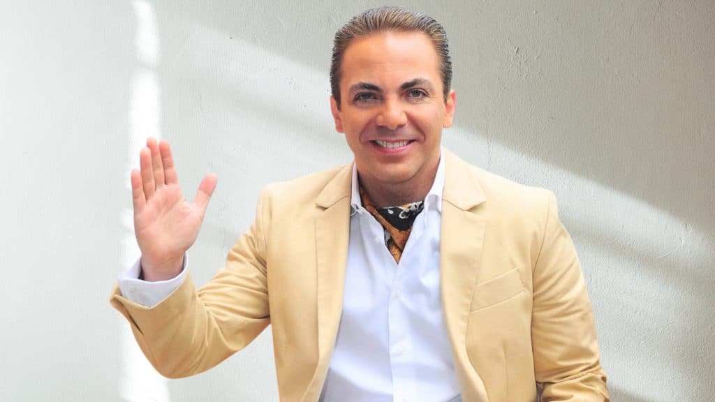 Cristian Castro saludando con la mano y sonriendo en retrato promocional con traje beige, mostrando el carisma del famoso cantante mexicano de baladas románticas