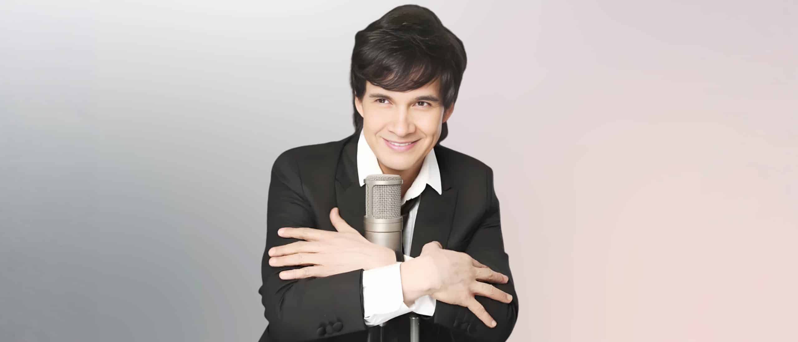 Gilberto Gless sonriendo frente a un micrófono de estudio, vestido con traje negro, en retrato promocional elegante para presentaciones en vivo y shows musicales