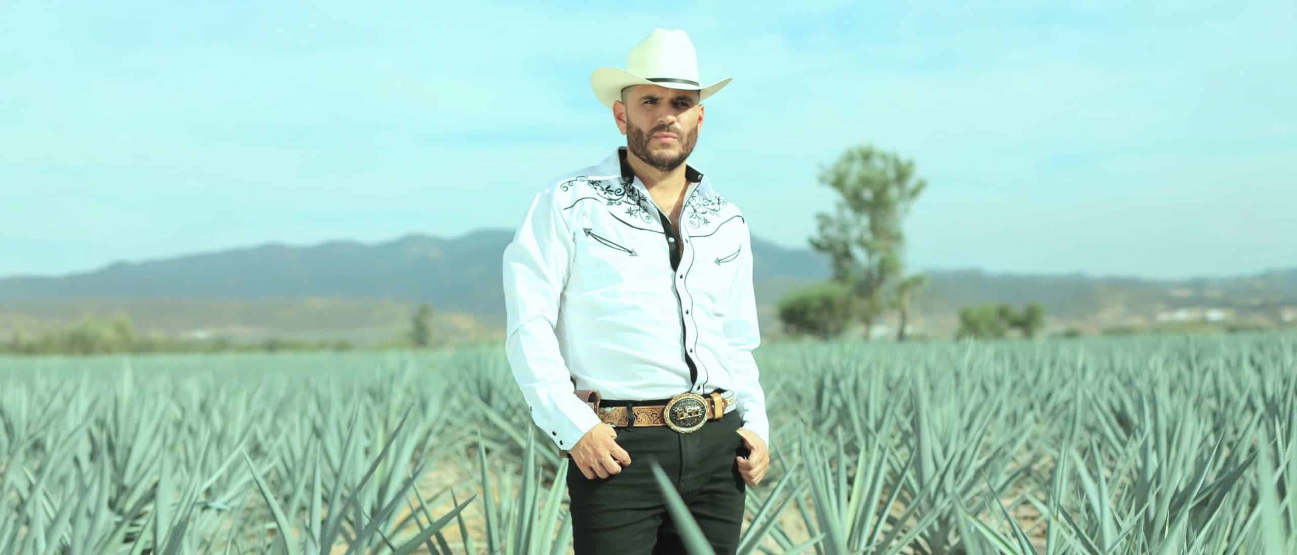 El Komander en locación campestre entre agaves con sombrero y camisa vaquera, imagen del regional mexicano con estilo auténtico y presencia imponente