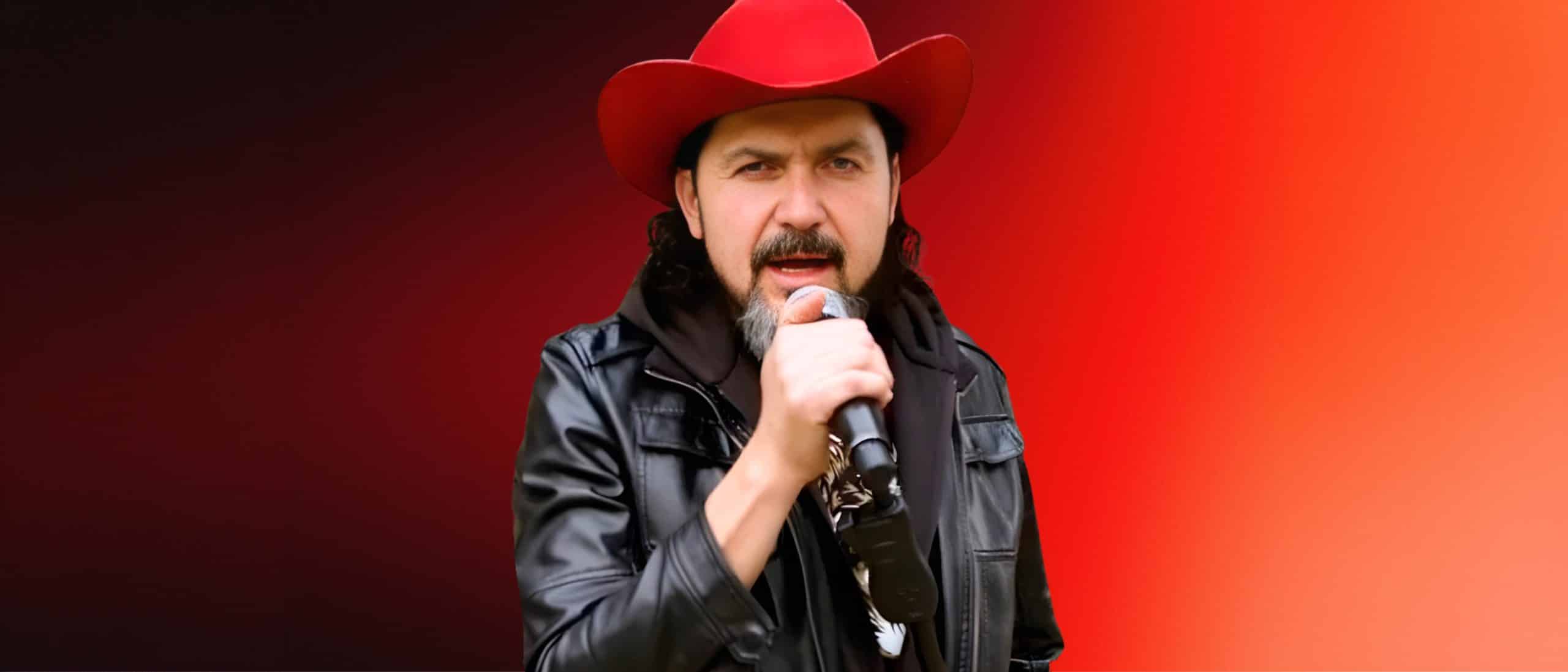 Reyli Barba cantando en vivo con sombrero rojo y chamarra negra, sosteniendo micrófono en primer plano sobre fondo rojo intenso en concierto enérgico