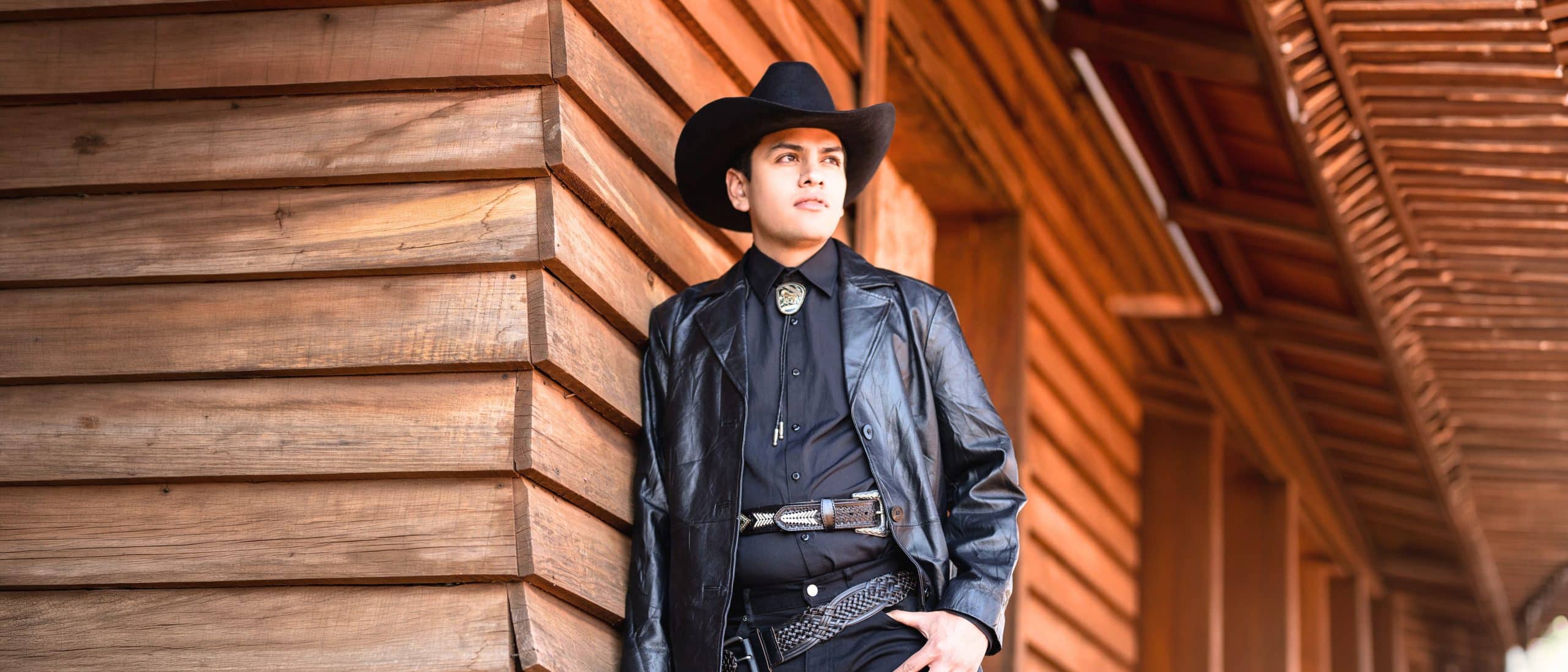 Virlán García en look regional con sombrero y chamarra de piel, posando con estilo frente a construcción de madera, ideal para promoción y contratación